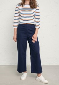 Gestreepte longsleeve top met blauwe, oranje en witte strepen, gecombineerd met wijde marineblauwe pantalons en witte sneakers op een gestructureerde achtergrond.