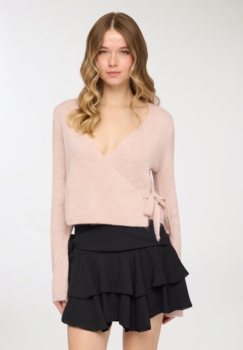 Maglione avvolgente in cashmere rosa chiaro con scollatura a V profonda e dettaglio a fiocco laterale, abbinato a una minigonna nera con balze. Texture morbida e design casual.