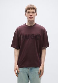 Vinrød oversized t-shirt med "HUGO"-logo i en mørkere vinrød farve, rund halsudskæring og korte ærmer. Båret med lyseblå jeans.