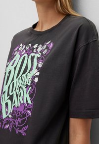 Osoba v tmavě šedém tričku s mátově zeleným a fialovým květinovým grafickým textem, na kterém je nápis "BOSS OWING THE DARK."