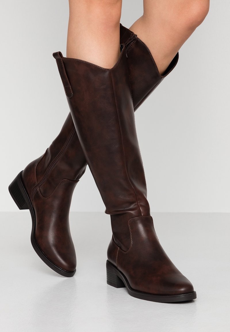 Bottes de cowboy en cuir brun jusqu'aux jambes avec talons empilés bas, portées sur des jambes croisées sur un fond clair uni.