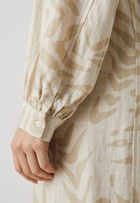 Chemise en lin beige avec un motif abstrait marron clair. Présente une manche froncée et un poignet à boutons pour le détail de la fermeture.