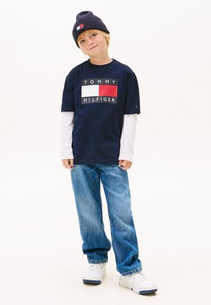 Tricou de bumbac bleumarin cu logo alb și roșu, purtat sub o cămașă albă cu mâneci lungi. O pereche de blugi albaștri din denim și adidași albi completează ținuta.