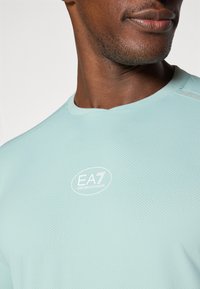 Svetlo modra mrežasta športna majica s krožnim izrezom, z vezenim logotipom EA7 Emporio Armani na prsih.