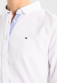 Camisa de algodón blanca con acentos de color azul claro en el cuello, cierre de botones y pequeño logotipo bordado en el pecho. Presenta una textura en el tejido.