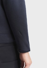 Manica di giacca in pile blu marina con una texture morbida, caratterizzata da un logo ricamato sul polsino, abbinata a un indumento scuro a strati sottostante.