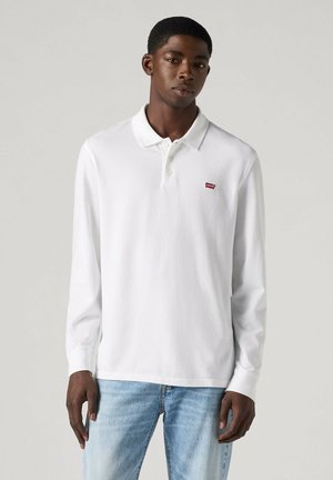 Młody mężczyzna w białej koszulce polo z długim rękawem z małym czerwonym logo Levi's i jasnoniebieskich dżinsach, stojący na jednolitym tle.