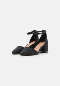Black suede heels with a pointed toe, ankle strap, and block heel. Features a nude insole and contrasting gold accents on the heel.