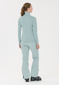 Vihreä, pitkähihaineen hiihtopaita, jossa on korkea kaulus, yhdistettynä yhteensopiviin hiihtohousuihin; sileä tekstuuri ja muotoonompelu. Mustat hiihtokengät täydentävät asua.