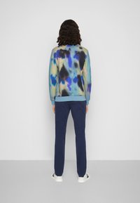 Felpa multicolore con motivo tie-dye nei toni di blu, verde e nero, abbinata a pantaloni blu navy e scarpe bianche, vista da dietro.