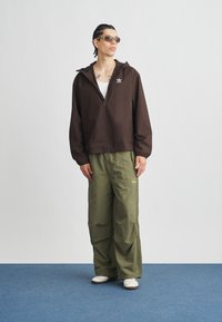 Καφέ ζακέτα με φερμουάρ, λευκό αμάνικο τοπ, άνετο χακί παντελόνι με λογότυπο, λευκά αθλητικά παπούτσια και γυαλιά ηλίου. Απλός σχεδιασμός, casual εφαρμογή, χαλαρή εμφάνιση.