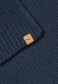 Marineblå strikket tekstil med et struktureret mønster og et lille lædertags med et logo, der er syet fast til stoffet.
