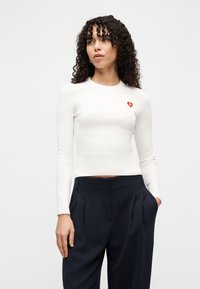 Femme aux cheveux bouclés portant un haut blanc à manches longues ajusté avec un logo cœur rouge et un pantalon noir ample à taille haute, les mains dans les poches.