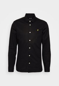 Camicia nera a maniche lunghe con bottoni bianchi e un piccolo logo giallo sul lato sinistro del petto, colletto piegato, posata su uno sfondo bianco.