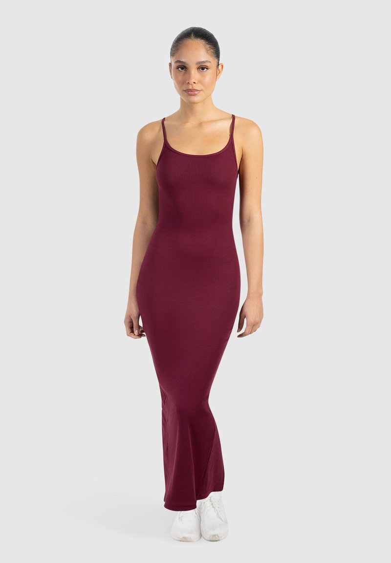 Kobieta stojąca przodem, ubrana w dopasowaną, bezrękawnikową, bordową sukienkę maxi oraz białe trampki na tle jednolitego, jasno-szarego tła.