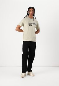 Camiseta de algodón verde claro con el logo negro de "Converse", combinada con pantalones negros y zapatillas beige. El modelo está de pie contra un fondo blanco.