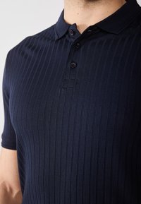 Tricou polo bleumarin cu textură textilă ribbed, guler cu trei butoane și mâneci scurte. Materialul este neted și are un luciu subtil.