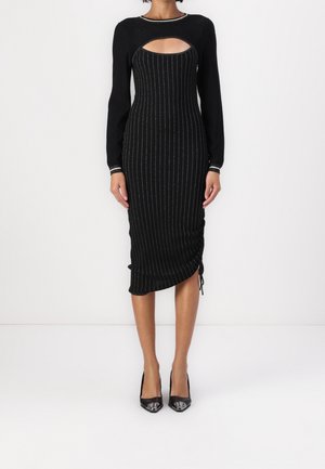 Donna che indossa un abito midi nero a maniche lunghe a righe sottili, con uno scollo sul petto e dettaglio arricciato sul lato, abbinato a décolleté nere a punta.