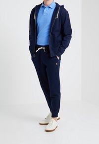 Marinblå zip-up hoodie med två framfickor över en ljusblå polo-skjorta. Mörka joggers med dragsko och vita sneakers.