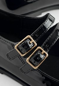 Černé lakované kožené boty s dvěma nastavitelnými popruhy se zlatými čtvercovými sponami. Design je elegantní s hladkým, lesklým povrchem.