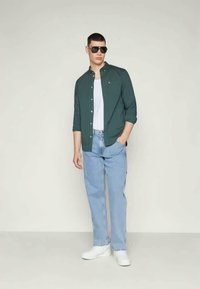 Tume roheline nupuga särk, valge T-särk selle all, heledad sinised teksad ja valged tossud. Modell kannab musti päikseprille, seistes neutraalse tausta ees.