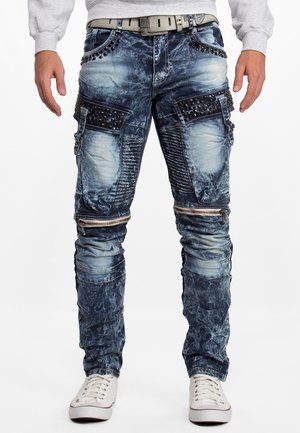 Man draagt acid-wash blauwe spijkerbroek met gespijkerde zakken, ritsen boven de knieën, geribbelde kniepaneel, beige riem, grijze trui en witte sneakers.