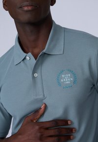 Gros plan sur une personne portant un polo bleu clair avec "MISE AU GREEN 1986" brodé sur la poitrine, la main reposant sur le polo.