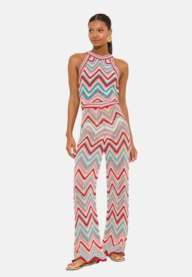 Woman standing wearing a sleeveless, multicolored chevron-patterned knit top and wide-leg pants with gold earrings and sandals.
