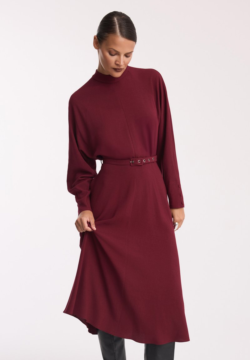Bordowa sukienka z długim rękawem, wysokim dekoltem, przewiązana w talii i z asymetrycznym dołem. Gładki materiał o subtelnej fakturze z guzikami na rękawach.