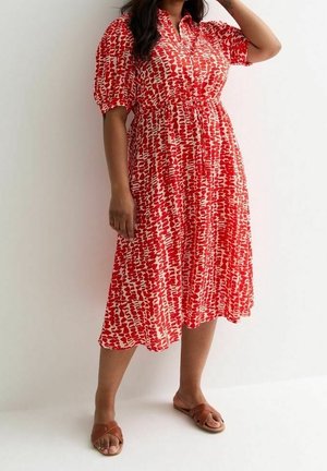 Frau trägt ein rot-weiß gemustertes Midikleid mit Puffärmeln und braunen flachen Sandalen und steht vor einer weißen Wand.