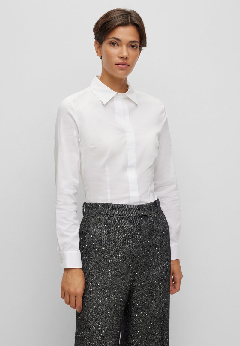 Camicia bianca con colletto e maniche lunghe, caratterizzata da una chiusura con bottoni frontale; abbinata a pantaloni neri a vita alta e testurizzati con puntini argentati.