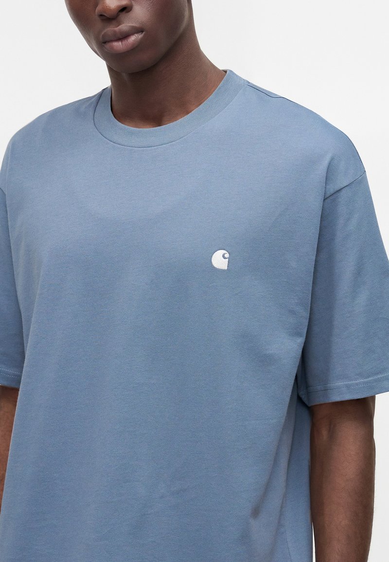Giovane uomo che indossa una t-shirt blu tinta unita a maniche corte con scollo a giro e un piccolo logo bianco sul petto sinistro.