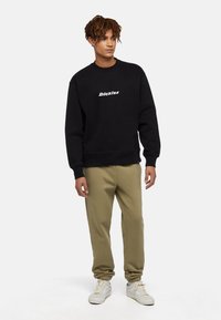 Zwarte sweatshirt met "Dickies" logo, rond de hals, en geribde manchetten, gecombineerd met olijfgroene joggers en witte sneakers. Glad weefseltextuur.