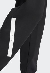 Černé tepláky s elastickými manžetami mají bílý vertikální pruh a text "SPORTSWEAR" na levé noze. Měkká, strukturovaná tkanina.
