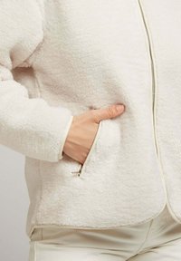 Veste en polaire crème avec fermeture éclair sur le devant, surface texturée et poches latérales. Présente des contours clairs contrastants le long des manches et de l'ourlet.