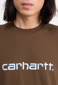 Hnedé bavlnené tričko s okrúhlym výstrihom, na prednej strane s logo "carhartt" v bielej farbe, umiestnené centrálne na hrudi. Hladká textúra, štandardný strih.