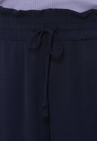 Ελαστική μέση με κορδόνι προσαρμογής σε μπλε σκούρες παντελόνες, φορεμένες με μοβ ριμπ μπλούζα.