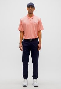 Άνδρας που φοράει κοραλί πόλο με κοντά μανίκια, ναυτικό μπλε παντελόνι με ζώνη, λευκά αθλητικά παπούτσια και σκούρο καπέλο μπέιζμπολ, στέκεται μπροστά σε απλό φόντο.