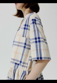 Vrouw draagt een crèmekleurige jurk met een blauw-beige ruitpatroon, voorzien van een groot borstvak en ellebooglange mouwen.