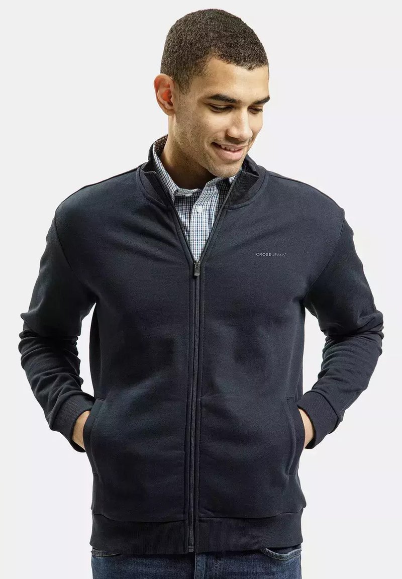 Granatowa bluza zapinana na zamek, wykonana z miękkiego materiału, z wysokim kołnierzem, bocznymi kieszeniami i ściągaczami na rękawach, noszona na koszuli w kratę.