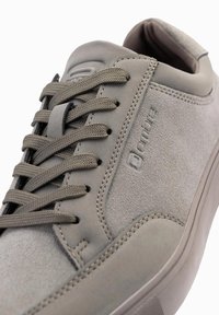 Šedé semišové tenisky s texturovaným svrškem, plochými šedými tkaničkami, polstrovaným límcem a nenápadným detaily loga na boku. Kulatá špička a gumová podrážka.