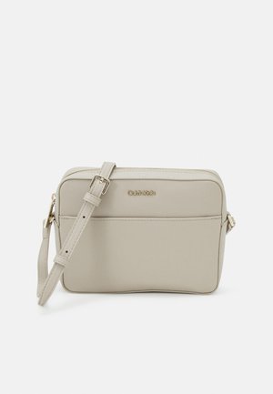 Sac bandoulière beige Calvin Klein avec poche avant, bandoulière réglable et logo doré sur fond blanc uni.