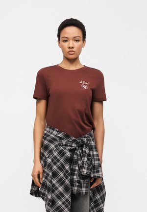 Ung kvinde iført en brun "le Soleil" T-shirt og en sort-hvid ternet skjorte bundet om livet, stående foran en ensfarvet baggrund.