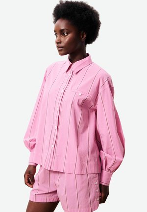 Vrouw met kort krullend haar, gekleed in een losvallend roze overhemd met verticale strepen en bijpassende shorts met knopen aan de zijkant.