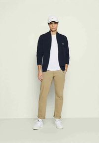 Tumed zip-up jakk, valge T-särgi, khaki pükste ja valgete tossudega. Aksessuaaride hulka kuulub valge müts väikese logoga.