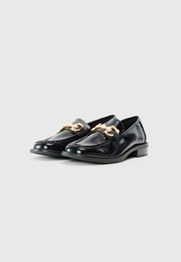 Mocasines de charol negro con un tacón bajo, que presentan un detalle decorativo de cadena dorada a lo largo del empeine y una superficie texturizada.