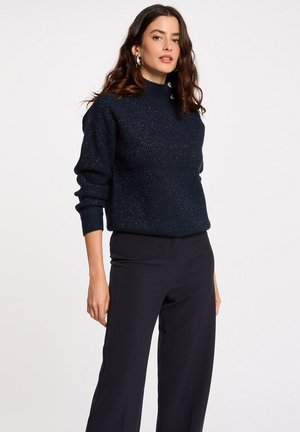 Pull à col haut en maille côtelée bleu marine, avec des fils métalliques subtils. Associé à un pantalon large noir.