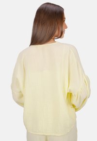 Locker sitzende gelbe Bluse aus leichtem Stoff mit strukturierter Oberfläche. Sie hat einen weiten Halsauschnitt und überschnittene Schultern, mit Knopfdetails.