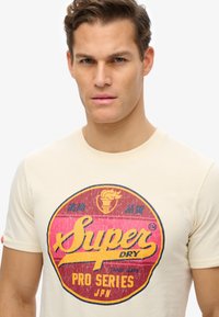 Camiseta de algodón color crema con un gran gráfico de estilo vintage en rojo, amarillo y azul, con textura desgastada y diseño circular.