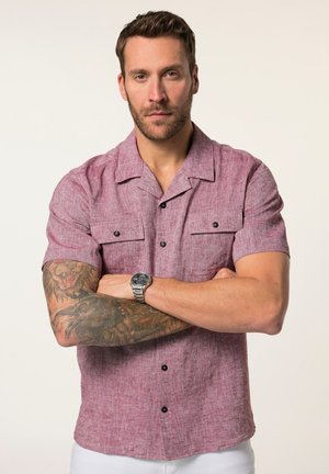 Uomo con capelli castani corti e barba, che indossa una camicia rosa a maniche corte con bottoni, mostrando un braccio sinistro tatuato in dettaglio e un orologio da polso.